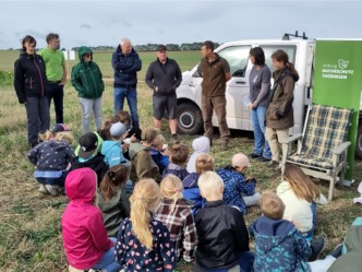 Mehrere Personen stehen vor sitzenden Kindern auf einer Wiese.