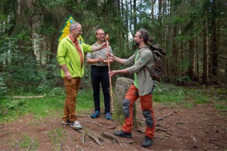 Drei Personen stehen im Wald. Eine Person übergibt einen Stock.