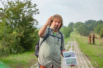 Ein Mann steht auf einem Kolonnenweg. Er hält ein kleines Buch mit Bildern in der Hand.